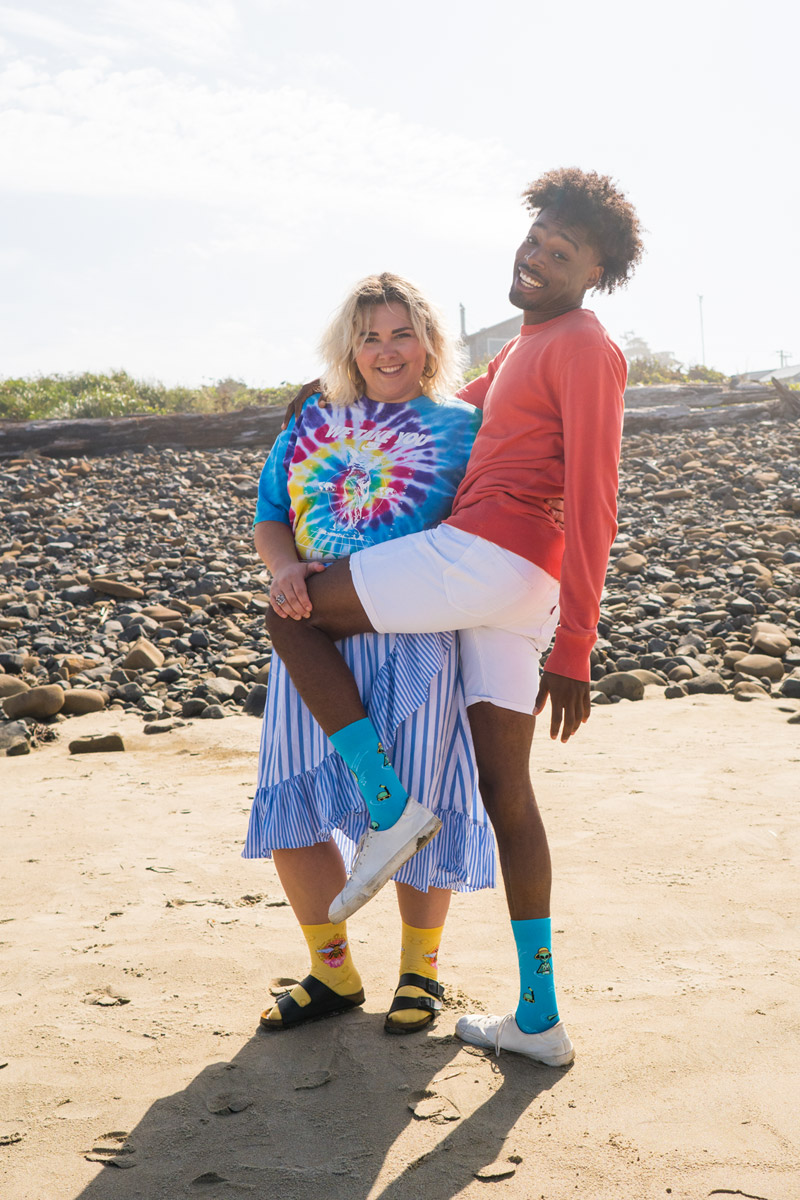 Two models standing on the beach while smiling. One model is holding the other models leg up by his knee.