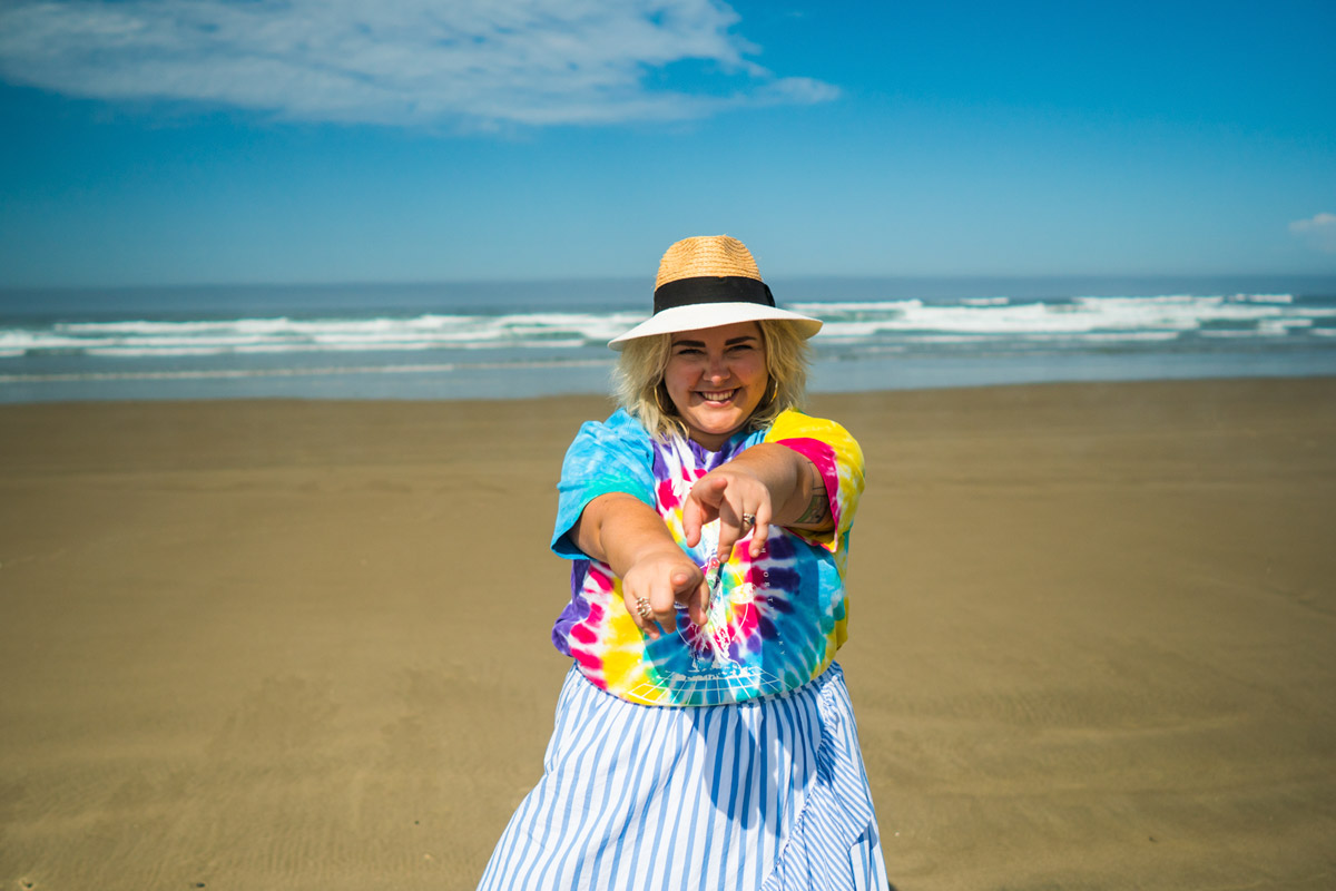 A woman model on a beach smiles while pointing both hands at the camera. The ocean is in the background.