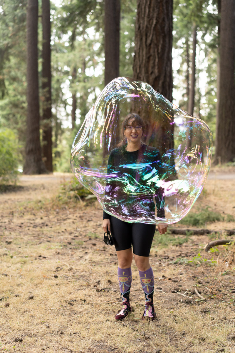 A model standing on a twig covered ground. Their face is obscured by a bubble half the size of her body.