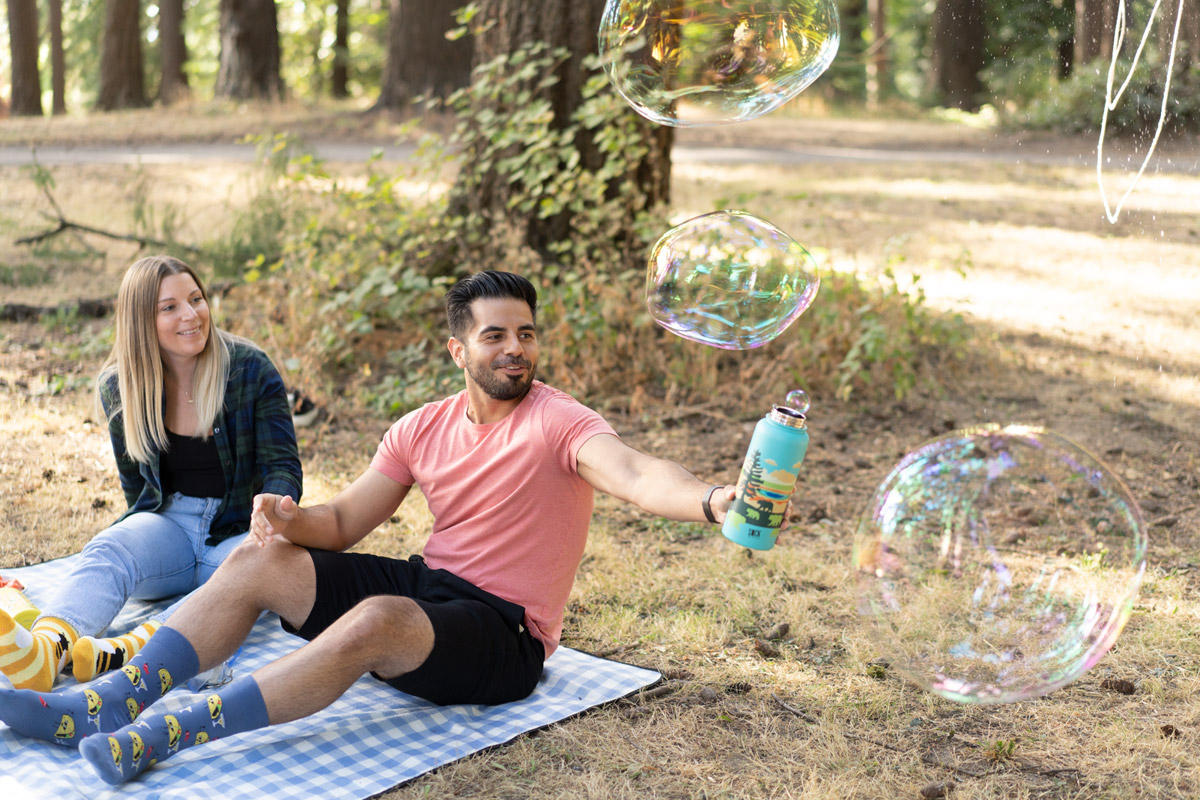 2 models sitting on a blue checkered blanket. One model is holding out a water bottle out to attempt to catch the many large bubbles that are floating by him.