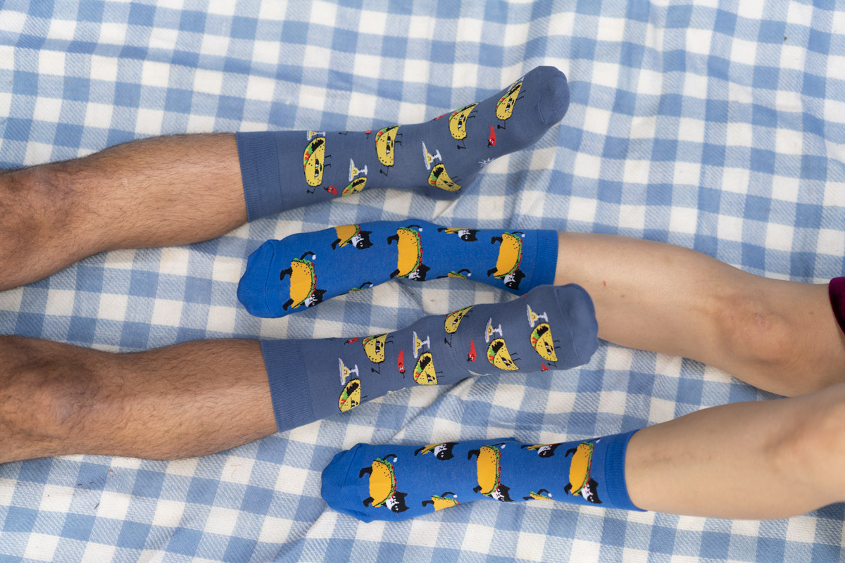 2 pairs of legs facing each other with their legs interlaced on a blue checkered blanket. They are each wearing different socks with tacos on them.