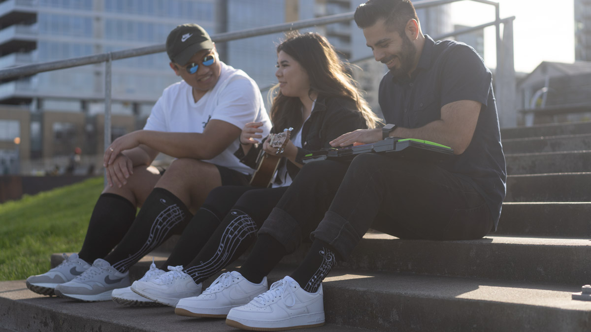 3 models sitting on a cement staircase outside. 2 models are smiling and chatting with each other, while the other is smiling while playing an electric keyboard.