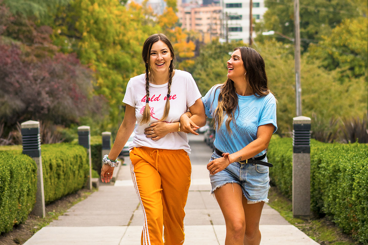 2 woman models are walking up a sidewalk with the Portland cityscape behind them. Their arms are linked and both are laughing.