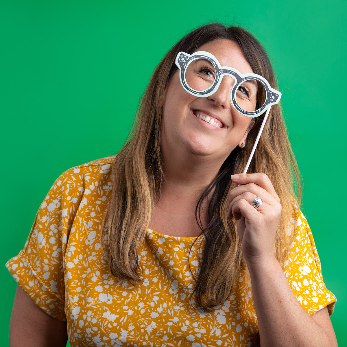 A woman smiling against a green backdrop, while holding up decorative prop glasses.