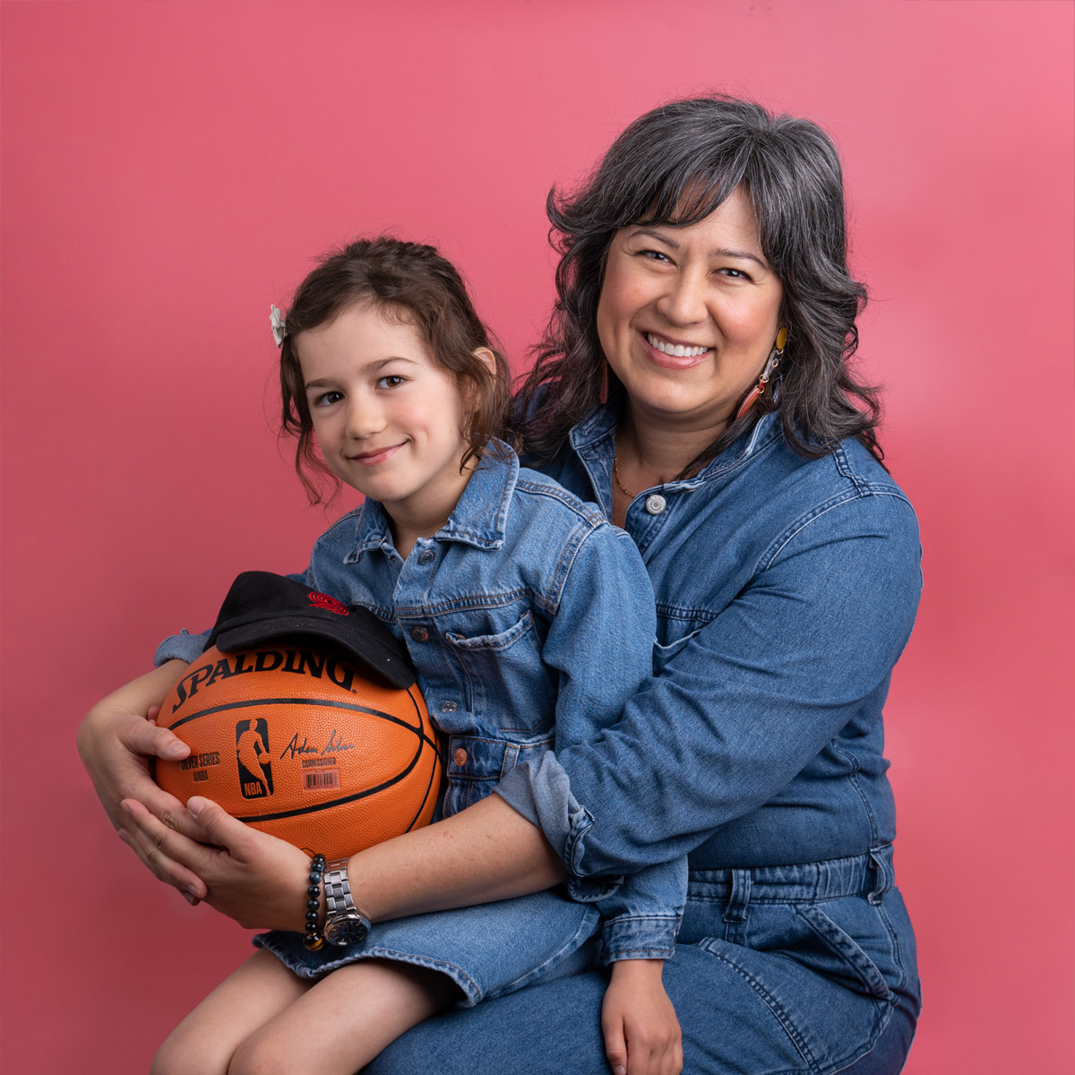 A child holding a basketball sitting in her mom's lap. Both are sitting in front of a pink backdrop.