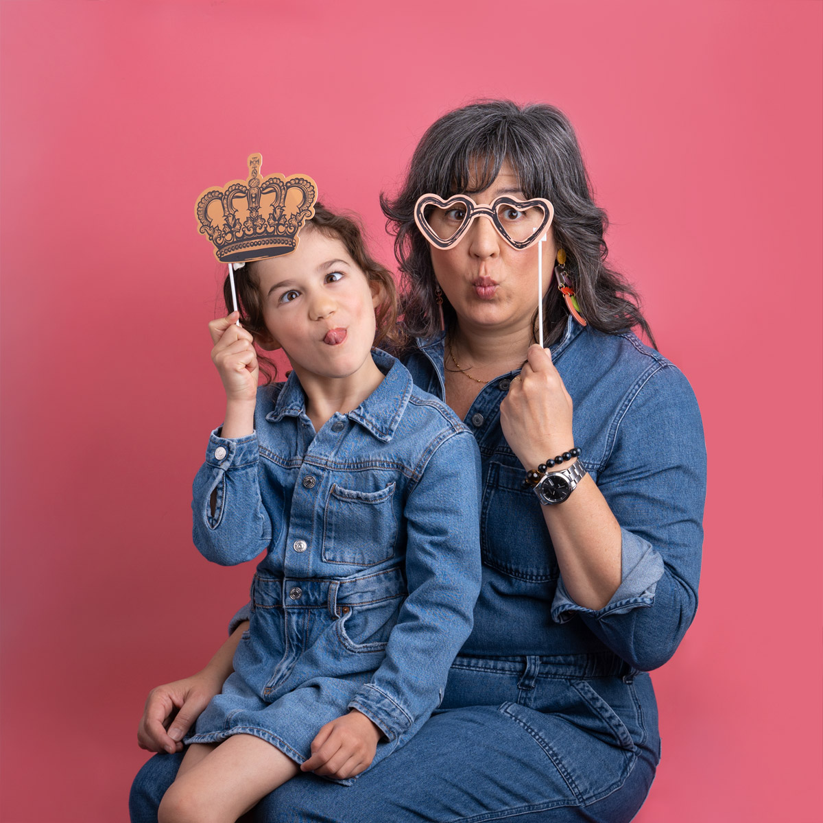 A child sitting in her mom's lap while they both make silly faces in front of a red backdrop.