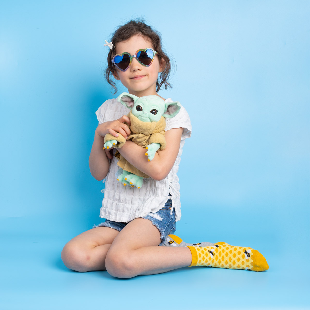 A child sitting on her knees while holding a Grogu doll and wearing blue heart shaped sunglasses.