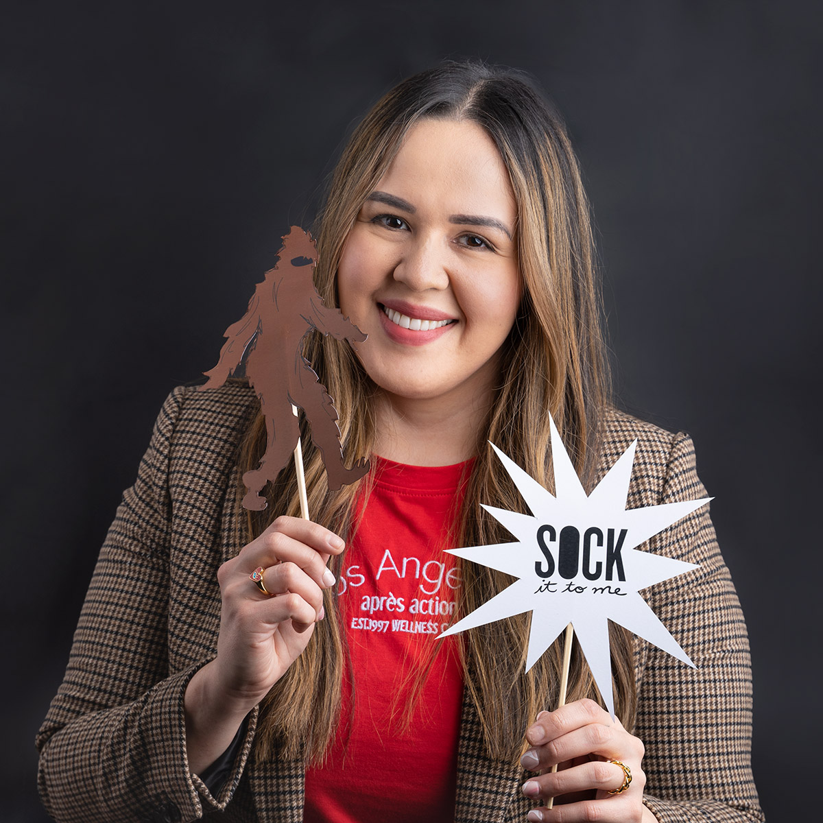 A woman sitting against a black backdrop, while holding up a sasquatch cut out and a Sock It to Me logo cutout.