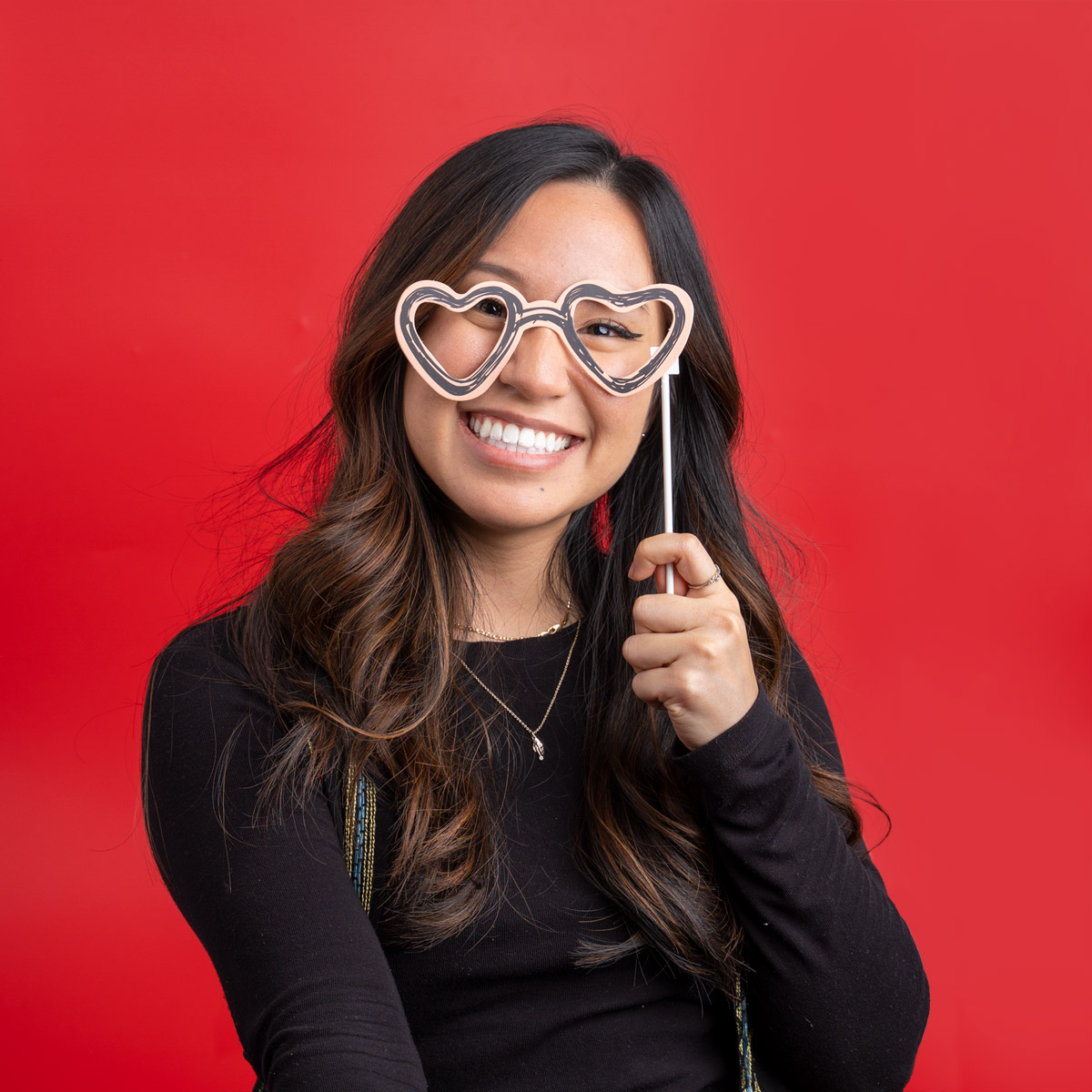 A woman smiling against a red backdrop, while holding up heart glasses.